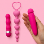 drie roze seksspeeltjes op een roze achtergrond, waaronder een hartvormig speeltje en een cilindervormig speeltje in een hand