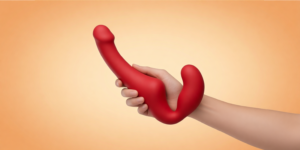 een rode dubbele dildo, vastgehouden door een hand tegen een oranje achtergrond