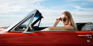 blonde vrouw in zonnebril zit in rode cabriolet met open dak aan de kust