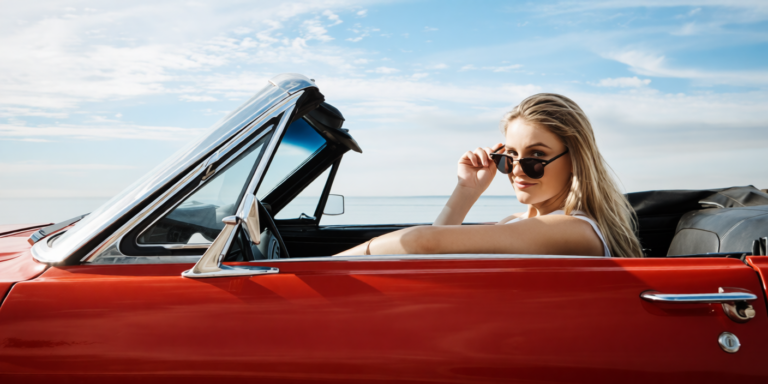blonde vrouw in zonnebril zit in rode cabriolet met open dak aan de kust
