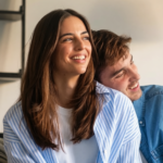 een vrouw met lang bruin haar glimlacht terwijl een man naast haar zit en ook glimlacht