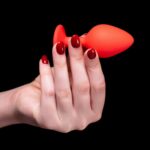 een hand met rode nagellak houdt een oranje plug vast tegen een zwarte achtergrond