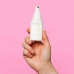 een hand houdt een witte sprayfles vast tegen een roze achtergrond