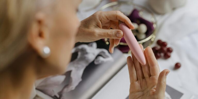 een vrouw houdt een roze vibrator vast terwijl ze naar haar hand kijkt, met druiven en een boek op de achtergrond