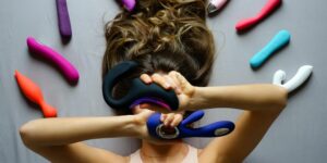 vrouw houdt twee vibrators vast terwijl ze op een grijze achtergrond ligt omringd door andere vibrators