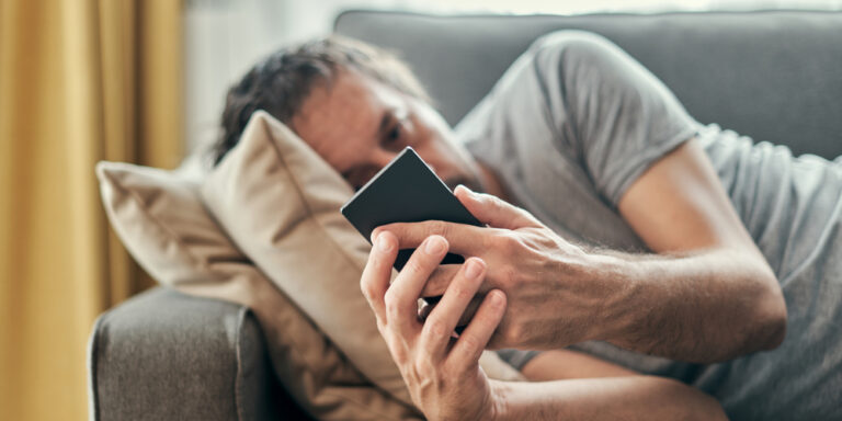 een man ligt op een bank en kijkt naar zijn telefoon