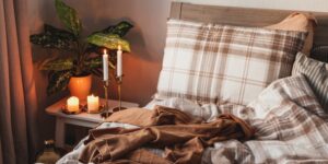 een bed met geruite lakens en een bruine deken, naast een nachtkastje met brandende kaarsen en een plant