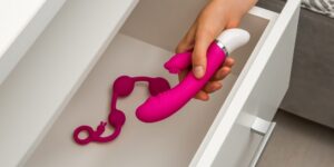 een hand houdt een roze vibrator vast terwijl een andere roze seksspeeltje op de grond ligt