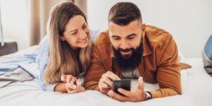 een glimlachende vrouw en een baardige man liggen op hun buik op een bed en kijken samen naar een smartphone