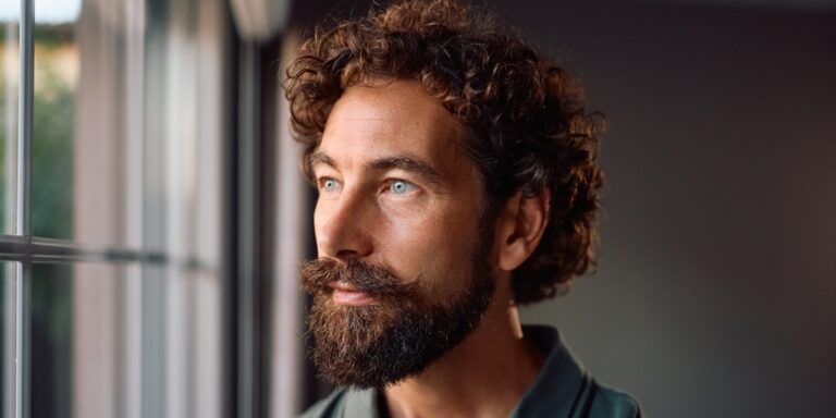 man met krullend haar en baard kijkt uit het raam
