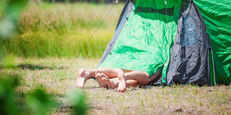 een persoon ligt met naakte benen buiten een groene en zwarte tent op een grasveld